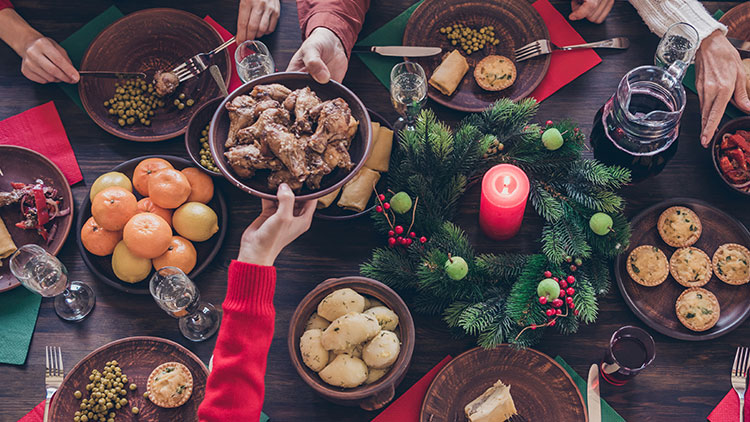 Festlich gedeckter Tisch mit grünem Adventskranz, roter Kerze, verschiedenen Speisen auf Tellern und mehreren Händen beim Servieren.
