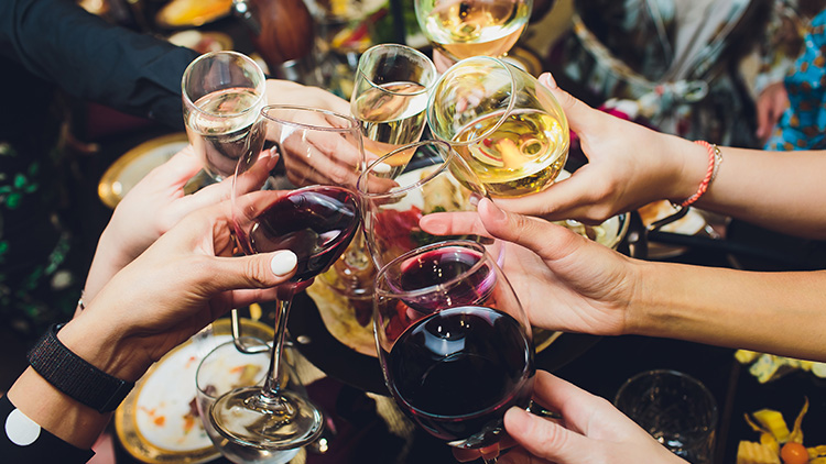 Mehrere Hände stoßen mit Weingläsern in verschiedenen Farben, darunter Rotwein und Weißwein, vor unscharfem Hintergrund an.