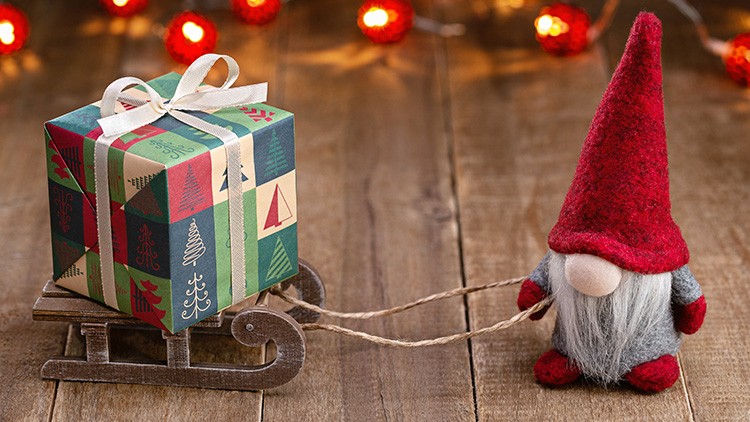 Ein kleiner Deko-Wichtel mit roter Mütze zieht einen Schlitten mit einem bunten, weihnachtlich verpackten Geschenk darauf.