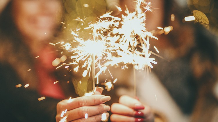 Zwei Hände halten leuchtende Wunderkerzen vor einem unscharfen Hintergrund mit warmem Bokeh-Lichteffekt.