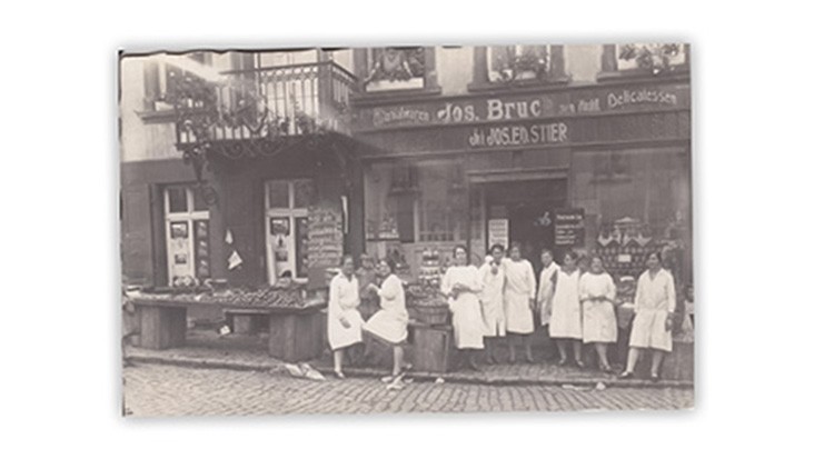 Eine historische Straßenszene zeigt ein Geschäft mit Schaufenstern, Markttischen und mehreren Personen in weißen Kitteln.