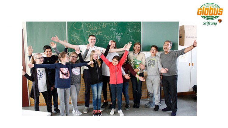 Eine Gruppe von Menschen steht vor einer grünen Tafel mit Schriftzügen wie „Danke“ und „GLOBUS“ und streckt die Arme nach oben.
