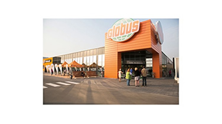 Eingang eines GLOBUS Marktes mit orangefarbener Fassade, Kunden und mehreren Pavillons mit Fahnen davor.