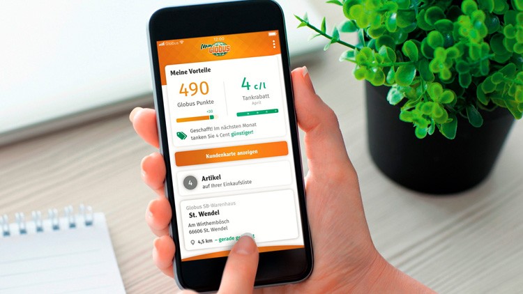 Eine Hand hält ein Smartphone mit der geöffneten mein GLOBUS-App, daneben eine grüne Pflanze und ein Notizbuch.