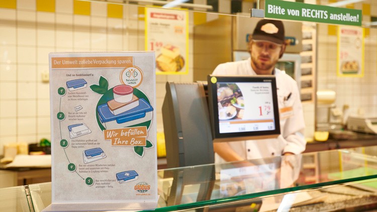 Plakat mit Text „Wir befüllen Ihre Box“ zeigt Mehrwegbehälter, dahinter Bedientheke mit Infobildschirm und Schild „Bitte von RECHTS anstellen!“.