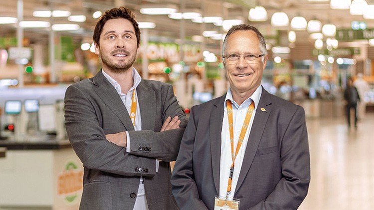 Zwei Personen in Businesskleidung mit orangefarbenen Lanyards stehen vor einer hell beleuchteten Supermarkt-Kulisse.