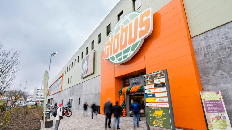 Eingangsbereich eines GLOBUS Markts mit orangefarbener Fassade, Globus-Logo, Eingangstür und Schildern, Menschen gehen hinein.