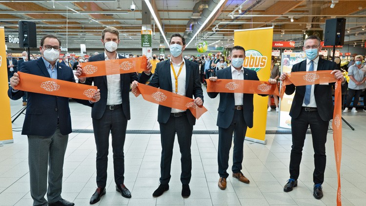 Fünf Personen in Anzügen halten ein oranges GLOBUS-Band vor einer Menschenmenge in einem Markt mit GLOBUS-Logo im Hintergrund.