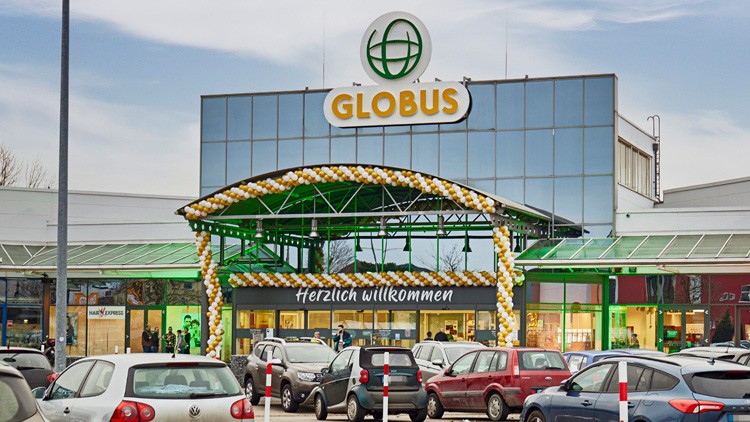 Eingangsbereich eines GLOBUS Marktes mit gelb-weißer Luftballondekoration und Schriftzug „Herzlich willkommen“, davor geparkte Autos.