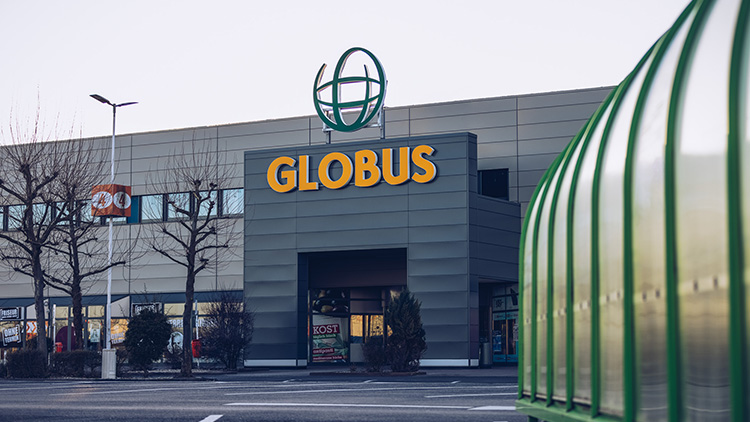 Das GLOBUS-Eingangsgebäude mit gelbem GLOBUS-Schriftzug und grünem Globini-Logo, Äste und Parkdach im Vordergrund.
