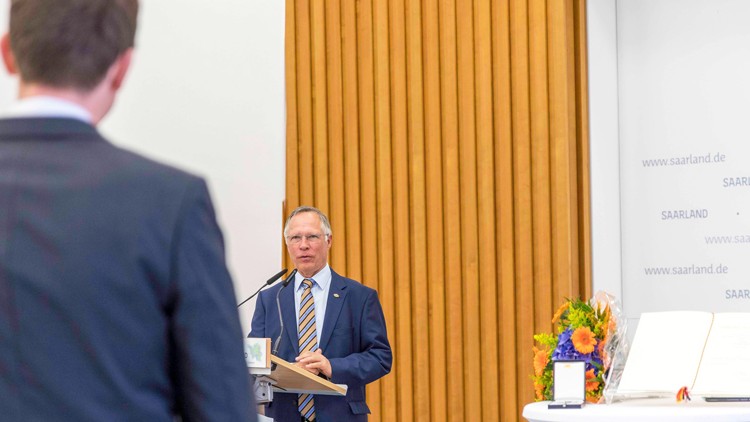 Ein Mann im Anzug spricht an einem Redepult, daneben ein Tisch mit Blumen, einem Buch und Beschriftung „saarland.de“.