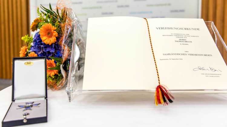 Auf einem Tisch liegen eine Verleihungsurkunde, ein Blumenstrauß und ein Etui mit einer Auszeichnung.
