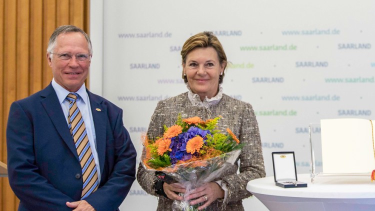Person mit Blumenstrauß steht neben einem Tisch mit Urkunde und Medaille, im Hintergrund mehrfach „Saarland“ auf Schriftwand.