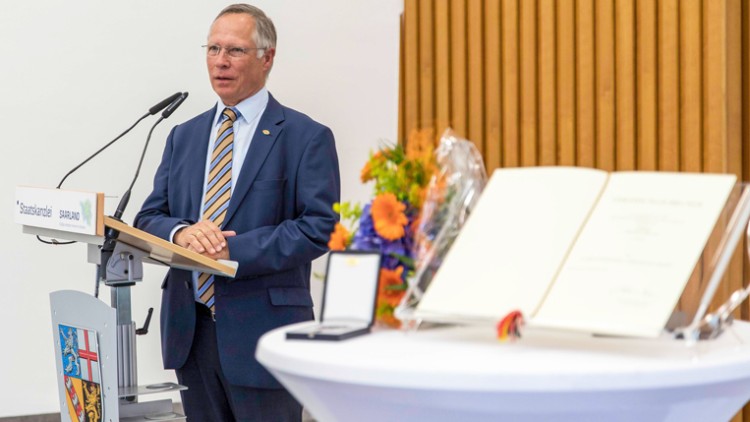 Ein Redner steht an einem Pult mit der Aufschrift „Staatskanzlei Saarland“, daneben ein Tisch mit Blumen und Dokumenten.