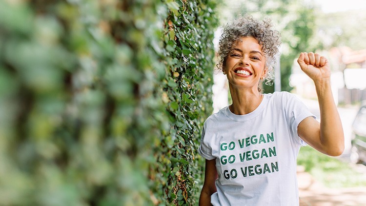 Person in weißem T-Shirt mit grünem Aufdruck „GO VEGAN GO VEGAN GO VEGAN“ steht vor einer blättrigen Wand und hebt eine Faust.