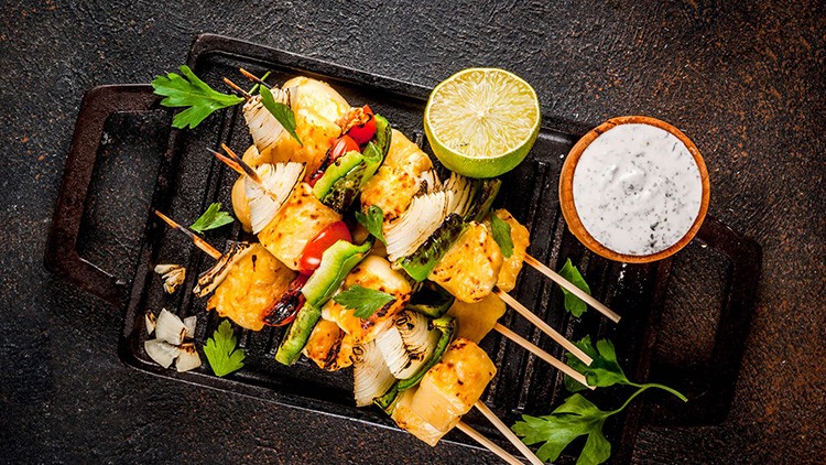 Gemüse-Käse-Spieße mit Paprika, Zwiebeln und Kräutern auf Grillpfanne, daneben Limettenhälfte und Schale mit weißem Dip.