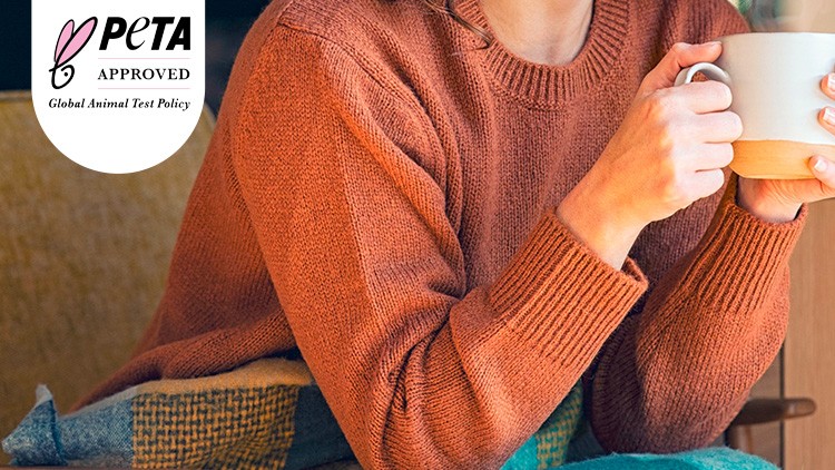 Eine Person in orangefarbenem Strickpullover hält eine weiße Tasse, oben links das PETA-Approved-Logo mit Text.