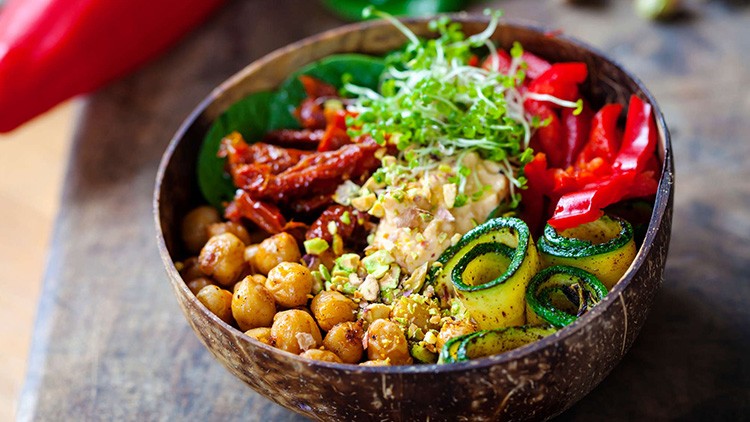 Bunte Bowl mit Kichererbsen, Zucchinischeiben, Paprika, Sprossen, getrockneten Tomaten, Hummus und frischem Blattgrün.