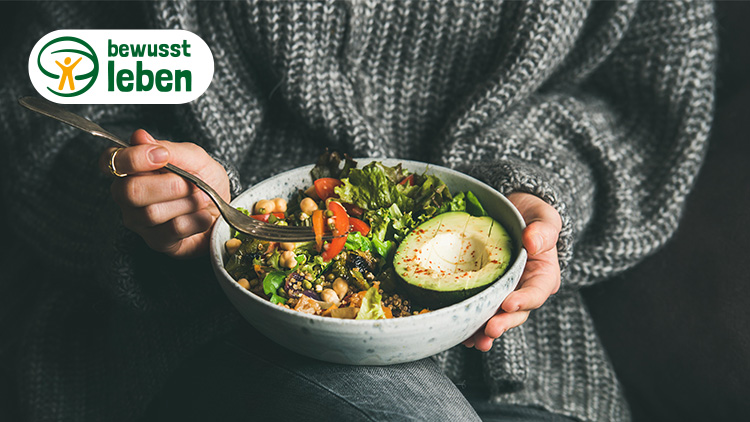 Hände mit grauem Pullover halten eine Schüssel mit Salat, Avocadohälfte und Kichererbsen, oben links steht „bewusst leben“.
