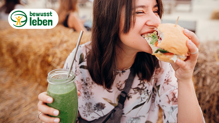 Person hält einen Burger in der Hand, daneben ein Strohballen und ein Glas mit Metallstrohhalm, Logo „bewusst leben“ oben links.
