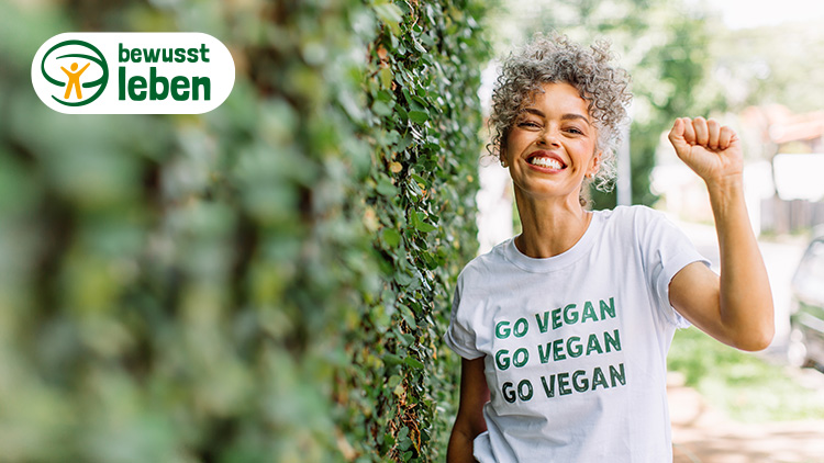 Person in weißem T-Shirt mit grüner Aufschrift „GO VEGAN GO VEGAN“ vor blätterbedeckter Wand, Logo „bewusst leben“ oben links.
