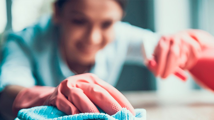 Person mit pinken Gummihandschuhen reinigt eine Oberfläche mit einem blauen Tuch, ein heller Hintergrund ist leicht unscharf.