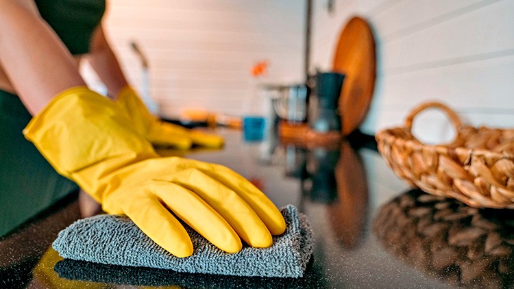 Hände mit gelben Gummihandschuhen wischen mit grauem Tuch eine glänzende Küchenarbeitsfläche ab, im Hintergrund Küchenutensilien.