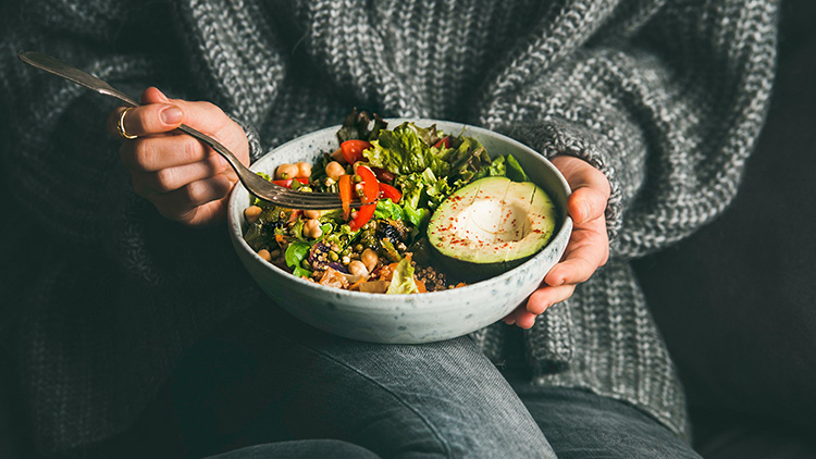 Eine Person hält eine Schüssel mit frischem Salat, Avocadohälfte, Tomaten, Kichererbsen und Quinoa, eine Gabel berührt den Salat.