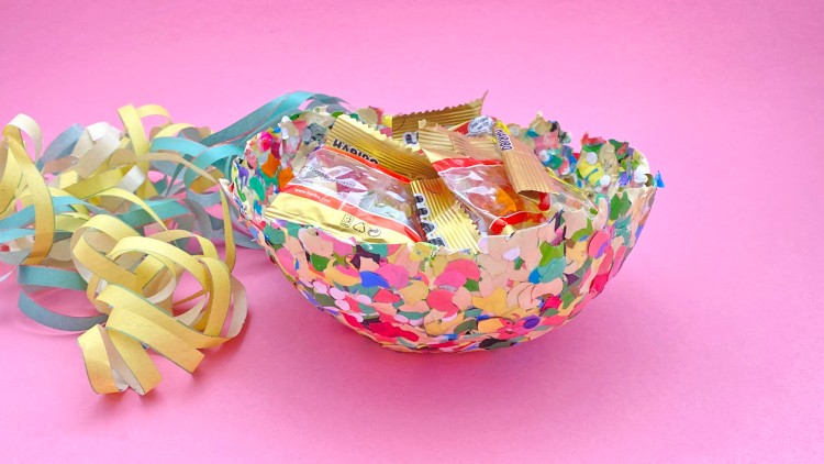 Bunte Schale aus Konfetti mit einzeln verpackten Gummibärchentüten, daneben gelb-blaue Papierluftschlangen auf rosa Hintergrund.