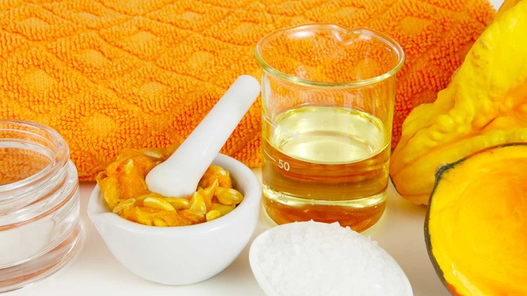 Ein Glas Becher mit Öl, ein weißer Mörser mit Kürbisstücken, grobes Salz und ein orangenes Handtuch liegen nebeneinander.