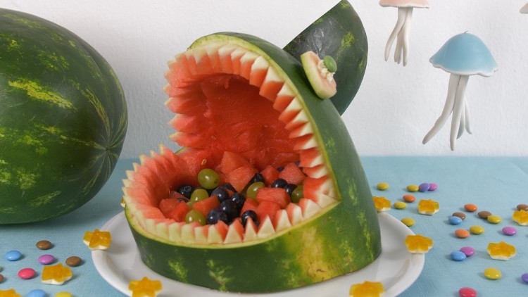 Eine ausgehöhlte Wassermelone mit gezackten Rändern, gefüllt mit Obst, steht auf einem Teller neben bunter Deko auf Tischdecke.