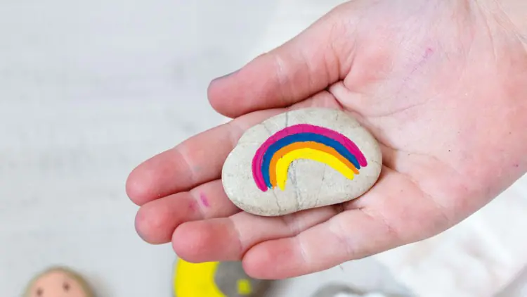Hand hält bemalten Stein mit buntem Regenbogen, weitere bemalte Steine mit Motiven wie Wolke und Pilz liegen auf hellem Untergrund.