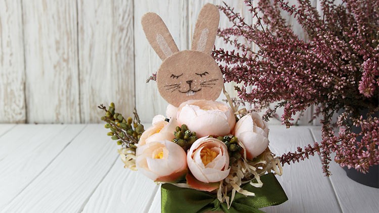Ein Blumengesteck mit rosa Pfingstrosen und grünem Band, dekoriert mit einer Hasenfigur aus Papier, steht auf weißem Holztisch.