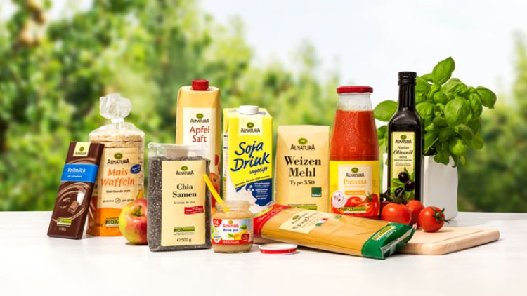 Verschiedene Alnatura-Produkte wie Saft, Öl, Mehl, Pasta und Gemüse vor unscharfem, grünem Gartenhintergrund arrangiert.