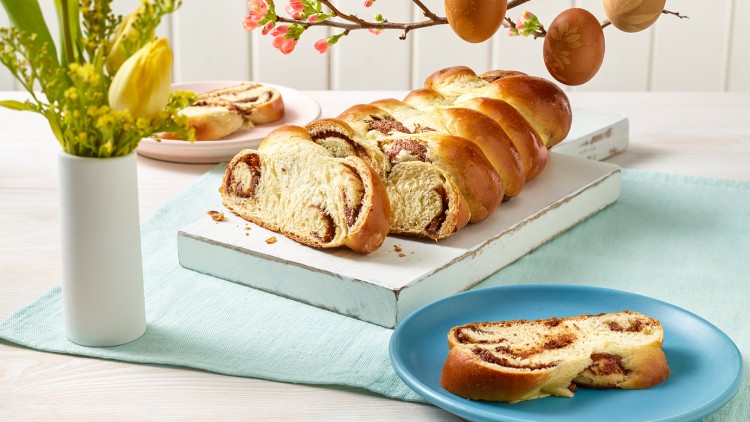 Ein angeschnittener Hefezopf mit Füllung auf einer weißen Platte, umgeben von Deko-Zweigen, Tulpen und bunten Tellern.