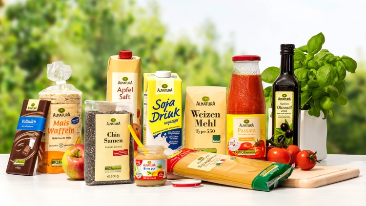 Eine Auswahl von Alnatura-Produkten wie Apfelsaft, Sojadrink, Passata und Schokolade steht auf einem weißen Tisch.