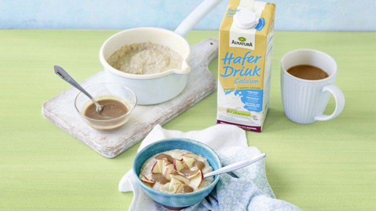 Eine Schale Haferbrei mit Apfelstücken, daneben Haferdrink-Packung, Kaffeetasse, kleiner Topf und Schale mit Soße auf grünem Tisch.