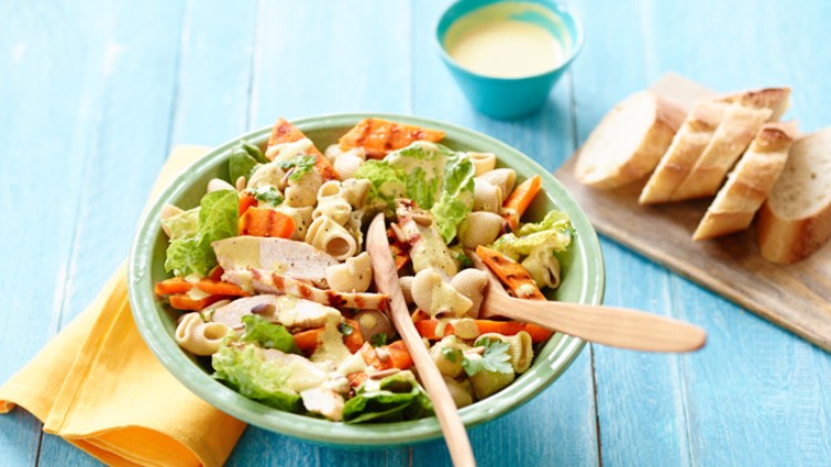 Grüner Teller mit Salat aus Hähnchen, Nudeln und Gemüse, daneben Baguettescheiben auf Holzbrett und gelbes Tuch.