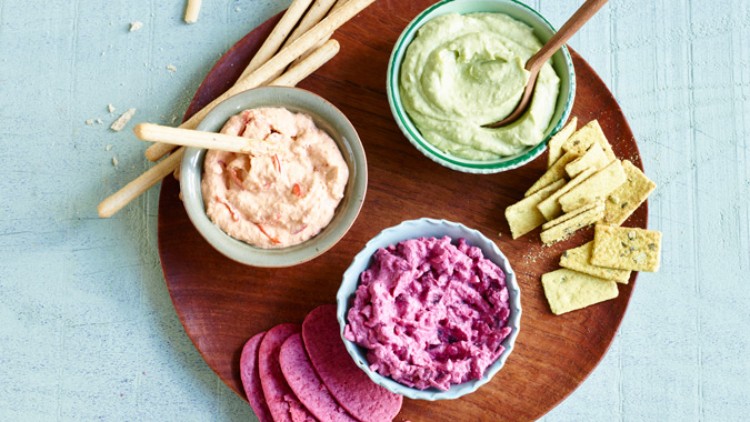 Drei Schalen mit Dips in Rosa, Grün und Lila auf einem Holzbrett mit Crackern, Brotsticks und rosa Aufschnitt.