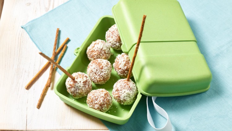 Grüne Brotdose mit kugelförmigen Snacks, bestreut mit Kokosraspeln und je einem eingesteckten Salzstangenstück, auf einem blauen Tuch.