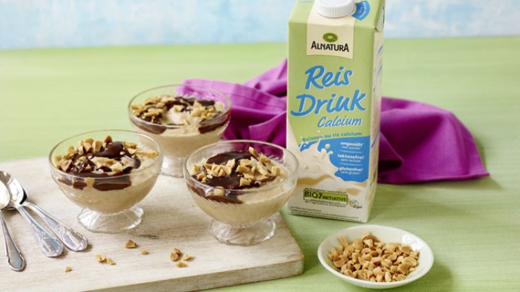 Drei Dessertschalen mit Reiscreme, darüber Schokosauce und Nüsse, daneben Alnatura Reis Drink Calcium und eine Schale gehackte Nüsse.