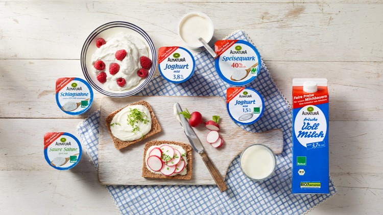 Verschiedene Alnatura-Molkereiprodukte, Brot mit Radieschen und ein Glas Milch auf einem Tisch mit blau-weiß karierter Unterlage.