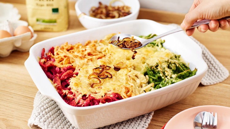 Weiße Auflaufform mit bunten Spätzle in Rot, Gelb und Grün, bestreut mit Käse und Röstzwiebeln, darauf eine Gabel mit Zwiebeln.