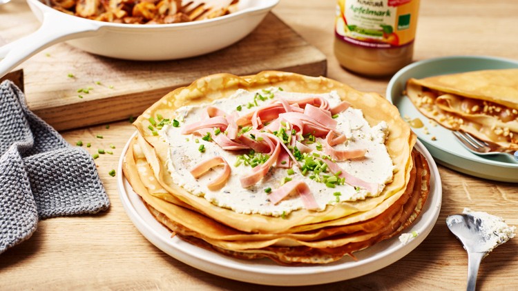 Ein Teller mit gestapelten Crêpes, belegt mit Frischkäse, Schinkenstreifen und Schnittlauch, steht auf einem Holztisch.
