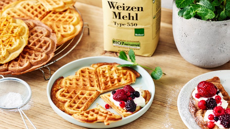 Frisch gebackene Herzwaffeln auf einem Teller mit Beeren und Sahne, daneben Weizenmehl Bio Type 550 und Minze in einem Topf.