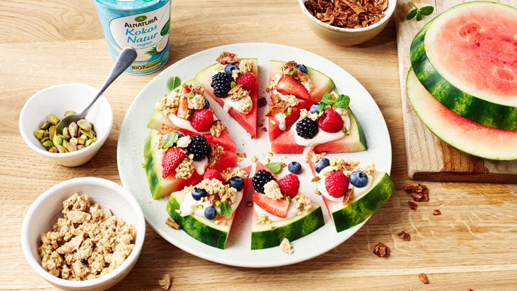 Melonenscheiben mit Joghurt, Beeren, Granola und Minze auf einem Teller, daneben Schalen mit Zutaten und ein Joghurtbecher.