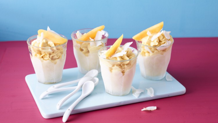 Vier Gläser mit einer Dessertcreme, Mango-Streifen und Kokosflocken auf weißem Brett, daneben vier weiße Löffel.