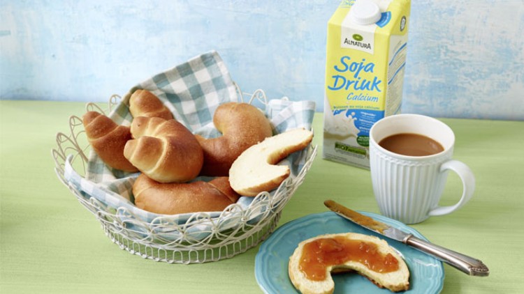 Ein Korb mit Croissants, eine Tasse Kaffee, ein Teller mit Marmeladenbrötchen und eine Packung Alnatura Soja Drink Calcium.