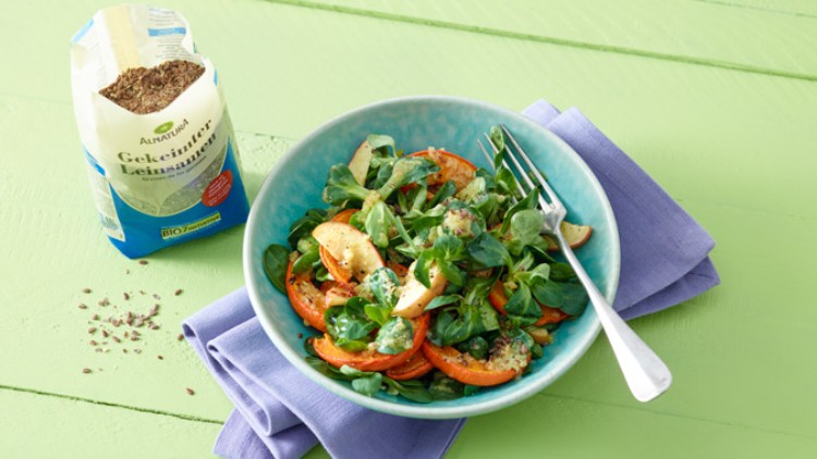 Ein grüner Salat mit Feldsalat, Apfelspalten, Karotten und Körnern in einer blauen Schale neben einer Packung Leinsamen.
