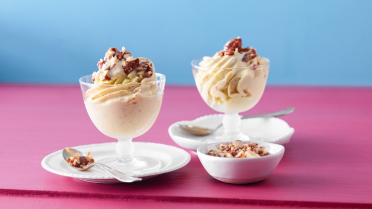Zwei Glasdessertschalen mit heller Eiscreme und Nüssen, daneben zwei Löffel und eine kleine Schale mit gehackten Nüssen.